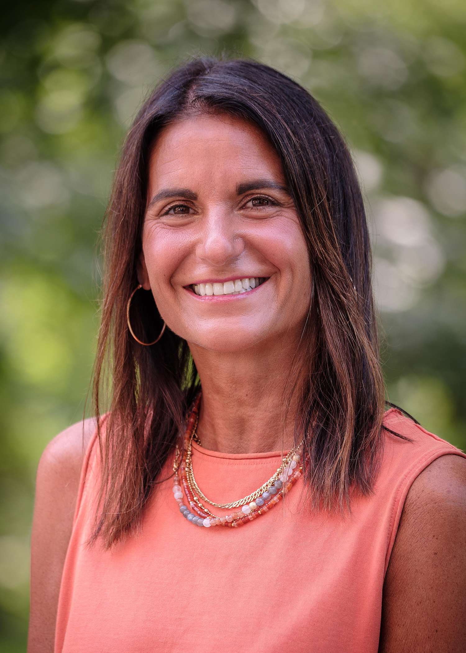 Shannon Milan Director of Admision wearing a sleeveless coral top and multiple layered necklaces stands against a blurred green outdoor background.