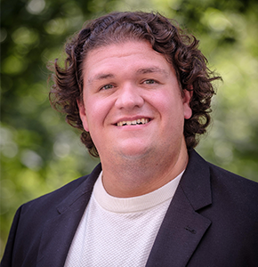 Beau Godkin, with curly dark hair and green eyes, poses outdoors against a lush green background. He wears a black suit jacket over a cream-colored knit turtleneck sweater.
