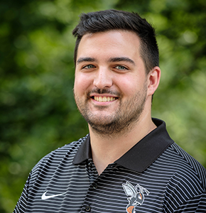 Douglas Maas with short dark hair, a trimmed beard, and blue eyes, poses outdoors against a lush green background. He wears a black and white striped Nike polo shirt featuring an orange and black hornet logo on the chest.