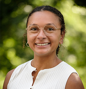Madison Ford, with curly dark hair tied back in a ponytail and gold hoop earrings, poses outdoors against a lush green background. She wears round gold-framed glasses and a sleeveless white button-up blouse.
