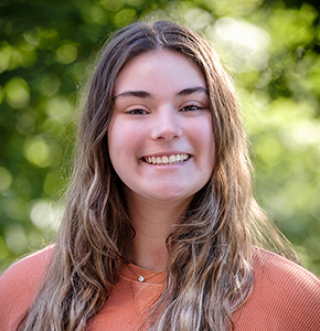 Jerrica Marietti, with long wavy light brown hair and almond-shaped brown eyes, poses outdoors against a lush green background. She wears a delicate pearl necklace over a sleeveless coral orange shirt.