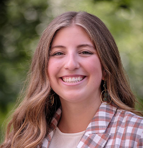 Sarah Pelyhes, with long wavy dark brown hair and brown eyes, poses outdoors against a lush green background. She wears dangling green hoop earrings and a brown-and-white checkered blazer over a white top.