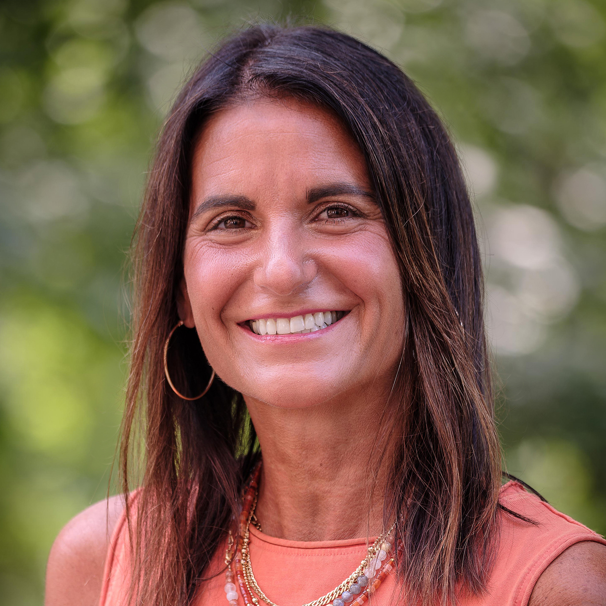 Shannon Milan, with shoulder-length dark brown hair and warm brown eyes, poses outdoors against a lush green background. She wears large gold hoop earrings and layered gold necklaces with bead accents over a sleeveless coral orange top.