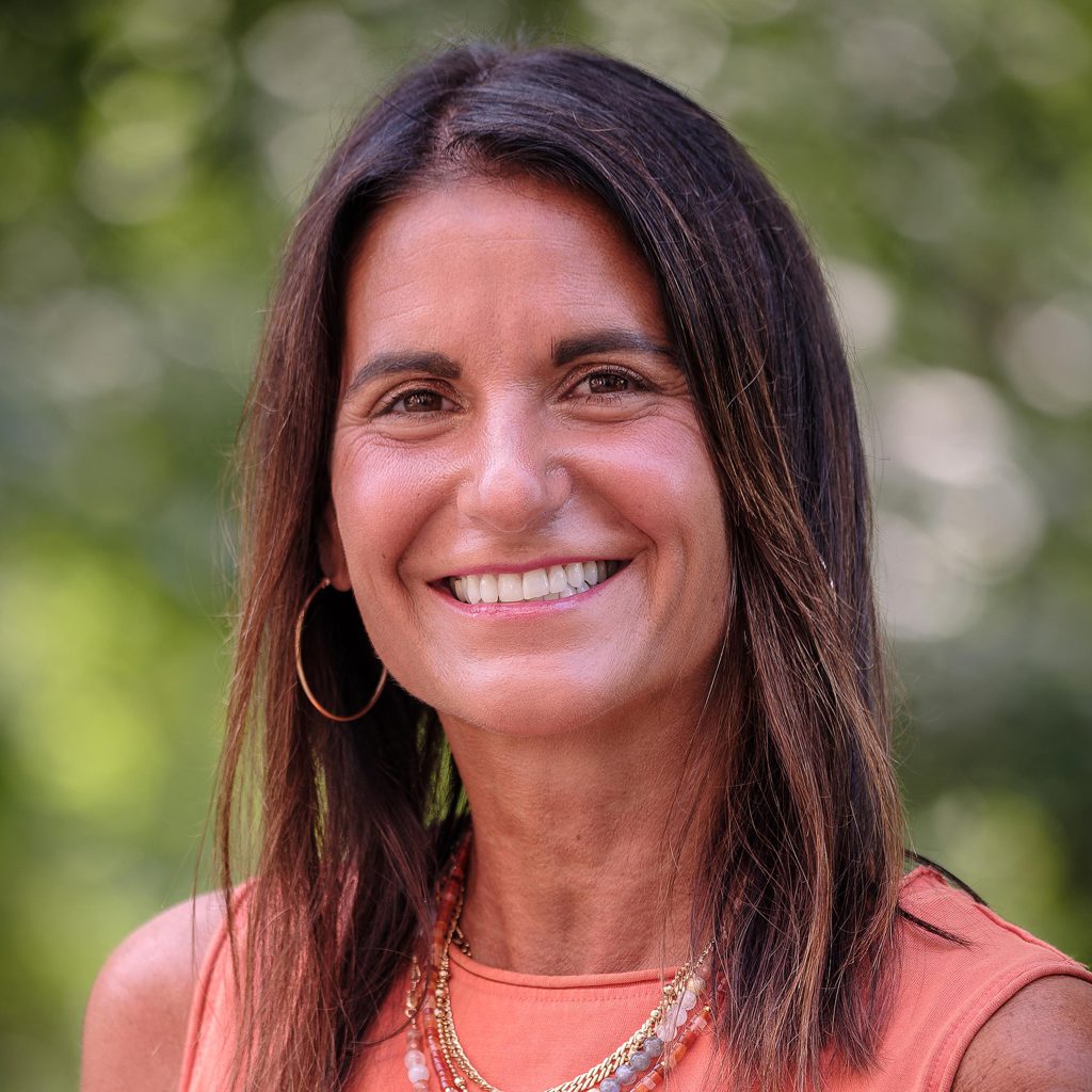 Shannon Milan, with shoulder-length dark brown hair and warm brown eyes, poses outdoors against a lush green background. She wears large gold hoop earrings and layered gold necklaces with bead accents over a sleeveless coral orange top.