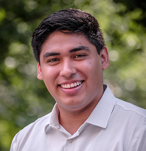 Victor M. Guerra Lopez, with short dark hair, warm brown eyes, and a smile, poses outdoors against a lush green background. He wears a short-sleeved white collared shirt.